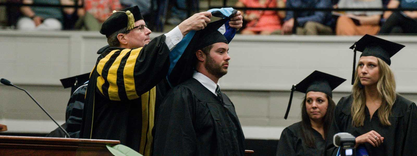 college dean put hoods around neck of graduate while other graduates watch and wait their turns